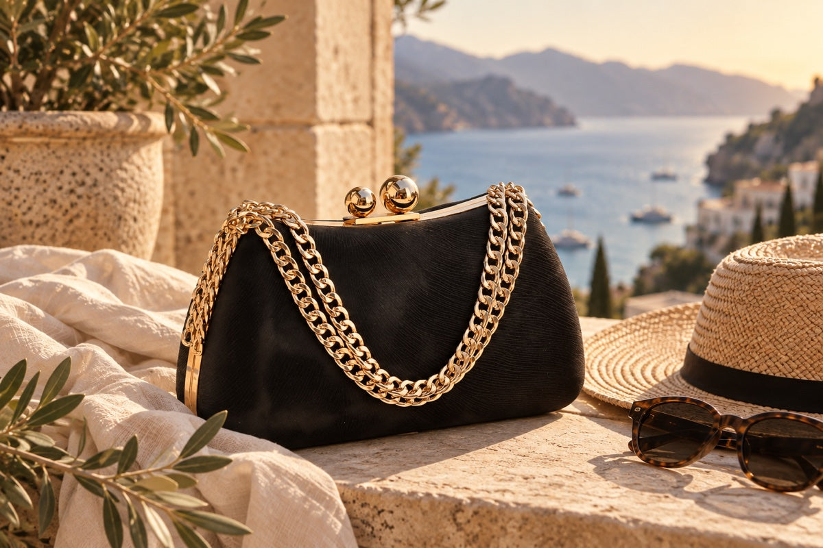 A Talia Guerrero clutch bag styled on a stone ledge with a straw hat, sunglasses and an olive branch, overlooking the coast.